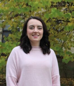 Sarah Dugan poses for a head shot photograph in front of a tree with green leaves.